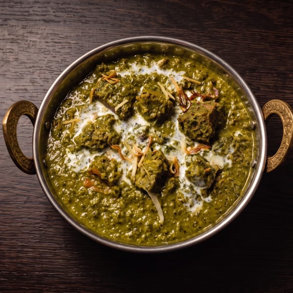 Indian kofta curry in creamy green sauce, garnished with cream and toasted slivers, served in an ornate handled bowl.