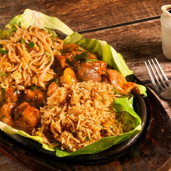 Steaming sizzling platter with noodles, fried rice, and a saucy main dish on lettuce, with cutlery.