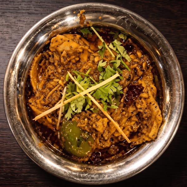 Overhead view of a richly spiced curry garnished with cilantro and ginger strips in a metal bowl on a dark wooden table.