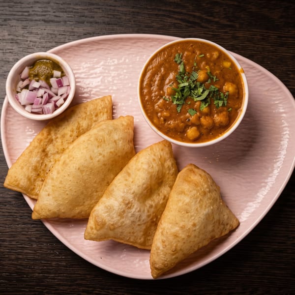 Golden samosas served with chickpea curry, chopped onions, and green chutney on a pink plate.