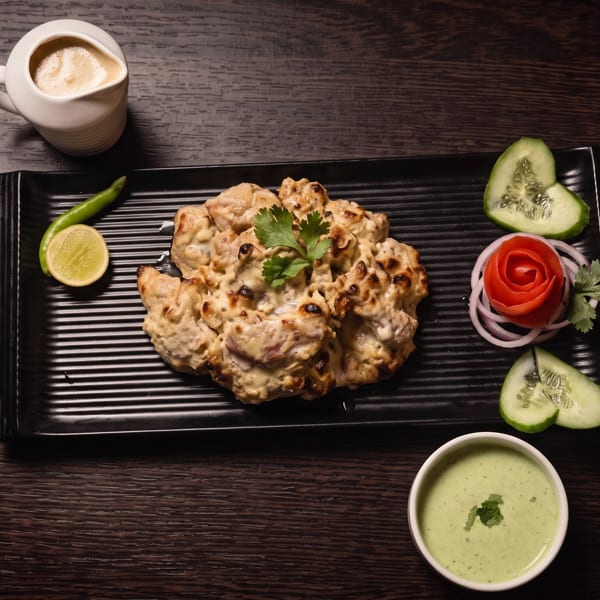 Grilled creamy chicken pieces garnished with cilantro, cucumber, tomato, onion, lime, chili, and green dipping sauce on a black platter.