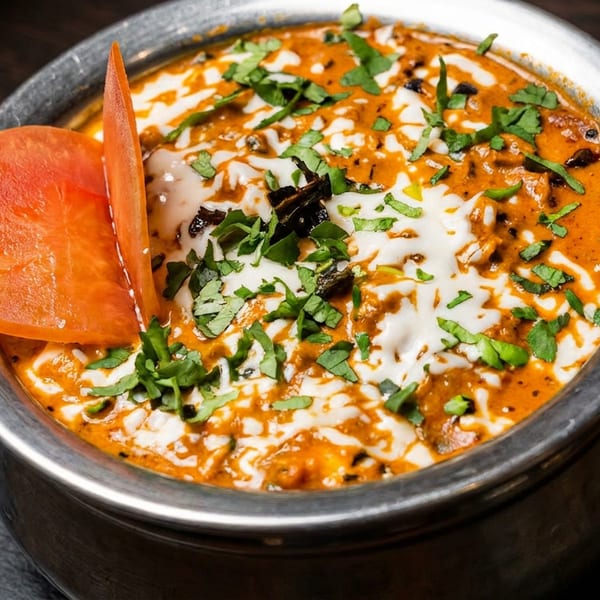 Indian curry with creamy orange sauce, cilantro, and a tomato garnish in a metal bowl.