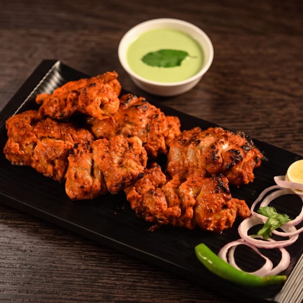 Plate of grilled orange chicken pieces with onion rings, lemon, green chili, cilantro, and mint sauce on a dark wooden table.