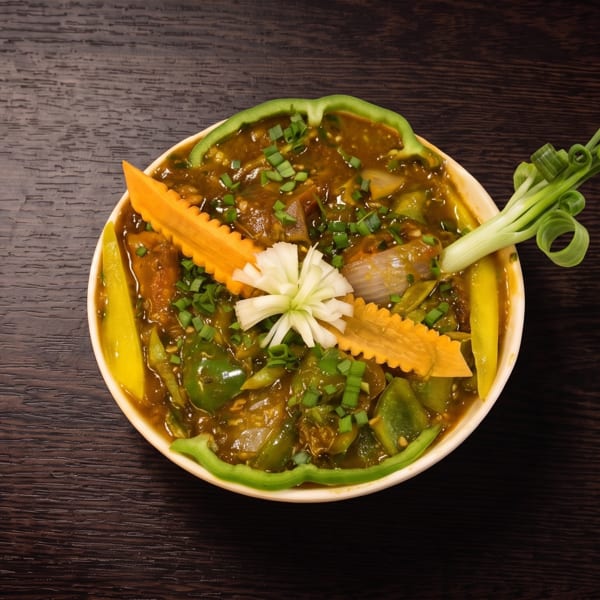 Bowl of glossy brown vegetable curry garnished with scallions, carved carrots, onion flower, and green pepper on a dark wooden table.