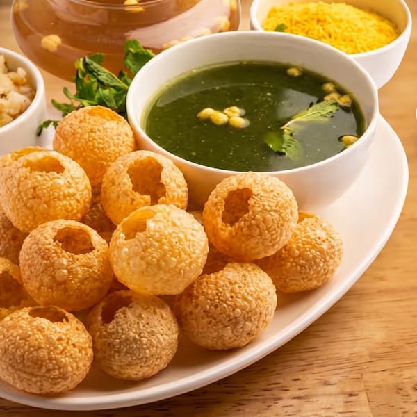 Crispy pani puri shells served with green spiced water, potatoes, yellow lentils, herbs, and chopped onions on a wooden table.
