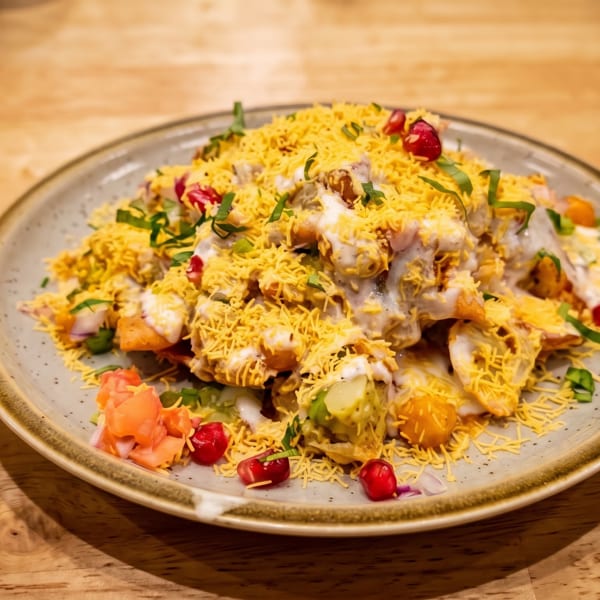 Indian chaat platter topped with sev, yogurt, herbs, pomegranate seeds, and colorful vegetables on a ceramic plate.