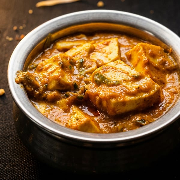 Vibrant paneer curry with cheese cubes in a metal bowl on a dark textured surface.