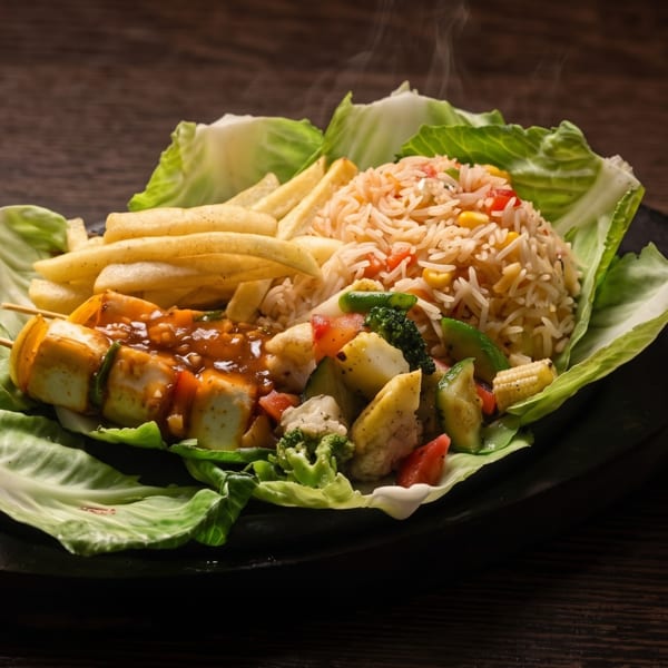 Steaming platter of vegetable skewers, fried potatoes, seasoned rice, and mixed vegetables on lettuce leaves.